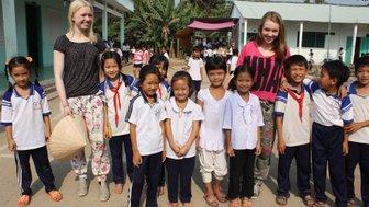 Zwei junge Frauen stehen lächelnd mit einer Gruppe von Kindern in Schuluniformen vor einer Schule im Mekong-Delta.
