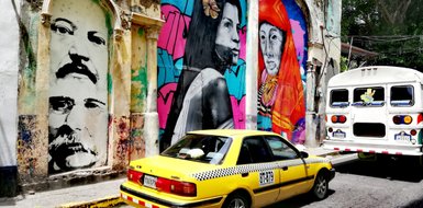 Bunte Wandmalereien zeigen Porträts von zwei Frauen und einem Mann vor einem hellgelben Taxi in einer urbanen Umgebung.