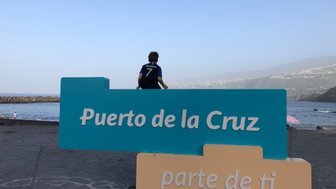 Ein Junge sitzt auf einem bunten Schild mit der Aufschrift 'Puerto de la Cruz' am Strand, während das Meer im Hintergrund glitzert.