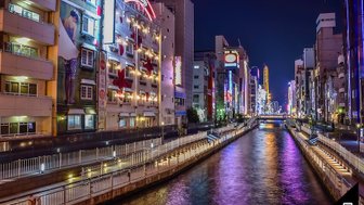 Die nächtliche Skyline von Osaka spiegelt sich im ruhigen Wasser eines Kanals, umgeben von bunten Neonlichtern und Gebäuden.