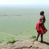 Ein Maasai in traditioneller Kleidung steht auf einem Felsen und blickt über die weite, grüne Landschaft der Masai Mara.