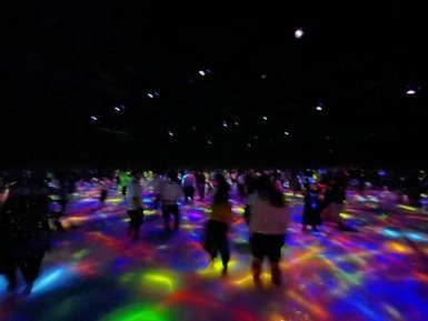 Eine große Menschenmenge bewegt sich über einen bunten, leuchtenden Boden in einem dunklen Raum voller Lichter.