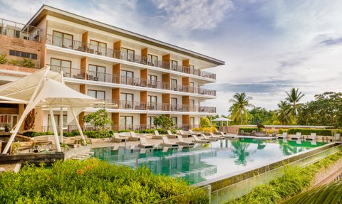 Ein modernes Resort mit mehreren Etagen, das von üppigem Grün und einem klaren Pool umgeben ist, unter einem strahlend blauen Himmel.