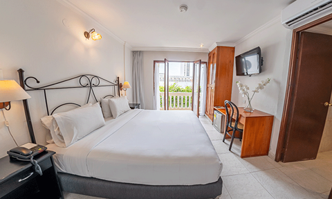 Ein helles Hotelzimmer mit einem großen Bett, einem Schreibtisch und einem Balkon, der auf einen grünen Außenbereich blickt.