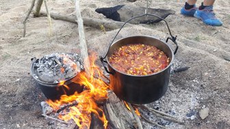 Ein großer Kochtopf mit buntem Eintopf kocht über einem offenen Feuer, umgeben von Sand und Holzstücken.