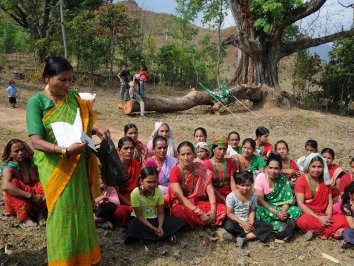 Eine Gruppe von Frauen in bunten Kleidern sitzt auf dem Boden und hört einer Rednerin aufmerksam zu.