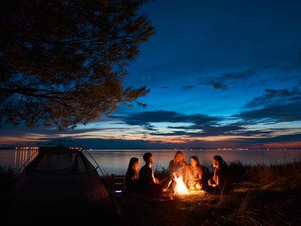 Eine Gruppe von fünf Freunden sitzt um ein Lagerfeuer am Ufer, während der Sonnenuntergang den Himmel in warmen Farben erleuchtet.