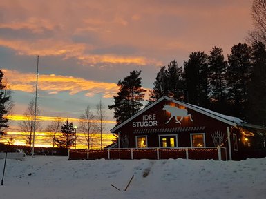 Ein rotes Holzhaus mit dem Schriftzug 'IDRE STUGOR' steht vor einem beeindruckenden Sonnenuntergang über schneebedecktem Boden.