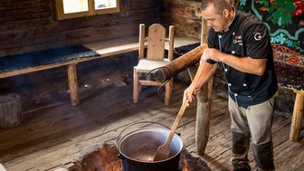Ein Koch rührt in einem großen Topf über einem offenen Feuer in einer rustikalen Holzhütte.