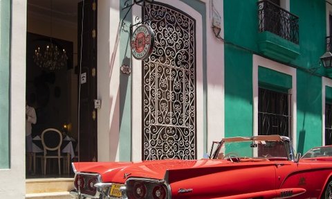 Ein leuchtend roter Oldtimer parkt vor einem eleganten Restaurant mit kunstvollem Eingang in Havanna.