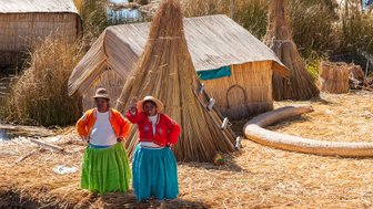 Zwei Frauen in traditioneller Kleidung stehen vor ihren Schilfhütten am Titicaca-See und lächeln freundlich in die Kamera.