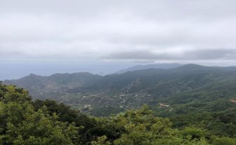 Eine weite Aussicht auf grüne Berge und Hügel, die unter einem bewölkten Himmel liegen, mit dem Ozean in der Ferne.