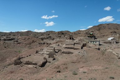 Eine weite, trockene Landschaft mit Überresten von alten Gebäuden, umgeben von sanften Hügeln und einem strahlend blauen Himmel.
