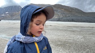 Ein Kind in einem blauen und gelben Regenjacke steht auf einem Gletscher, umgeben von grauen Wolken und Bergen.