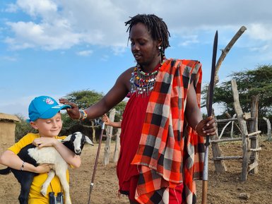 Ein Junge in einem gelben T-Shirt hält eine Ziege, während ein Maasai-Mann ihm freundlich den Kopf streichelt.