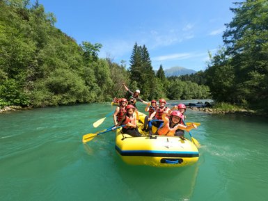 Eine Gruppe von Menschen paddelt fröhlich in einem gelben Raftingboot auf einem klaren, blauen Fluss, umgeben von üppigem Grün.