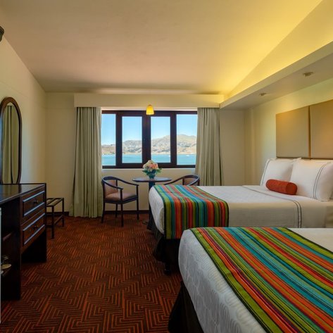 Ein helles Hotelzimmer mit zwei Betten, bunten Bettüberwürfen und Blick auf den See durch große Fenster.