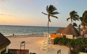 Ein ruhiger Strand mit sanften Wellen, umgeben von Palmen und traditionellen Strohdächern, während die Sonne untergeht.