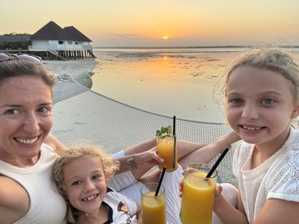Eine Frau und zwei Mädchen genießen fruchtige Cocktails am Strand, während die Sonne über dem Wasser untergeht.