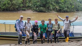 Eine Gruppe von sieben Personen posiert fröhlich am Ufer eines Flusses, umgeben von üppiger tropischer Vegetation.