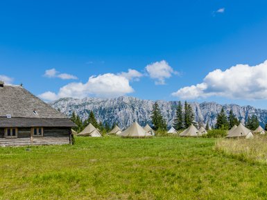 Eine rustikale Holzhütte steht im Vordergrund, umgeben von Zeltlagern und majestätischen Bergen unter einem strahlend blauen Himmel.