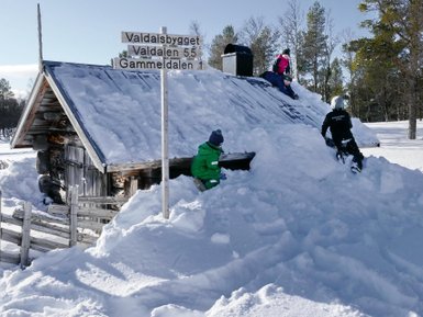 Zwei Kinder spielen im Schnee und klettern auf das Dach einer verschneiten Hütte, während ein Schild im Hintergrund steht.