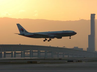 Ein Flugzeug landet majestätisch bei Sonnenuntergang über einer modernen Brücke und einer Stadtlandschaft im Hintergrund.