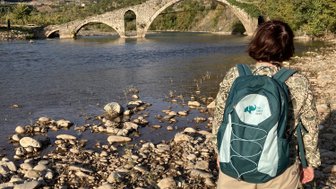 Eine Person steht am Ufer eines Flusses und betrachtet eine alte Steinbrücke, umgeben von grünen Hügeln.