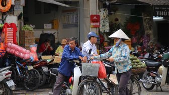 Eine Frau in traditioneller Kleidung verkauft frisches Obst auf einem belebten Markt in Hanoi, während Passanten vorbeigehen.