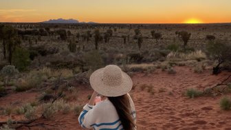 Eine Person mit einem Strohhut steht auf rotem Sand und genießt den Sonnenuntergang über der weiten, trockenen Landschaft.