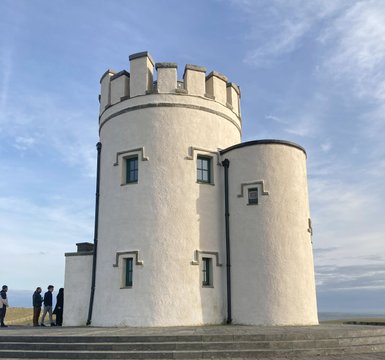 Ein historischer Turm mit runder Form und Zinnen steht vor einem klaren Himmel, während Besucher die Stufen hinaufgehen.