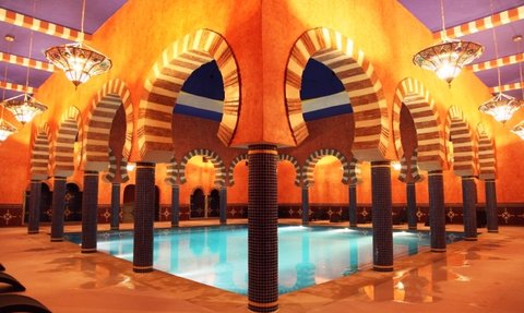 Ein eleganter Innenraum mit einem blauen Pool, umgeben von kunstvollen Bögen und warmen, orangefarbenen Wänden.
