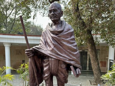 Eine bronzene Statue von Mahatma Gandhi steht auf einem Sockel, umgeben von Pflanzen und einem gepflasterten Weg.