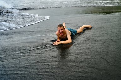 Ein fröhlicher Junge liegt im seichten Wasser am Strand, während die Wellen sanft über den schwarzen Sand rollen.