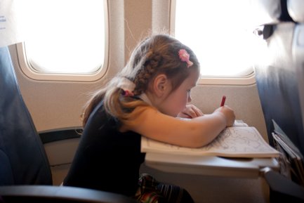 Ein kleines Mädchen sitzt am Fenster eines Flugzeugs und malt konzentriert in einem Malbuch mit einem roten Stift.