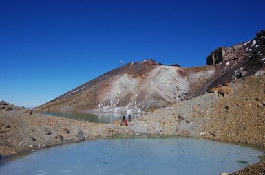 Ein klarer blauer Himmel über einer vulkanischen Landschaft mit einem gefrorenen Teich und schneebedeckten Bergen im Hintergrund.
