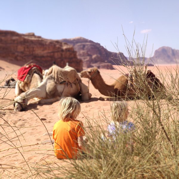 Zwei Kinder sitzen im Wadi Rum, während Kamele im Hintergrund auf dem Sand ruhen, umgeben von sanften Hügeln.