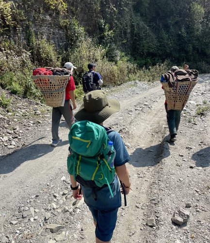 Drei Wanderer tragen große Körbe auf dem Rücken und gehen einen schmalen, steinigen Weg entlang, umgeben von üppiger Vegetation.