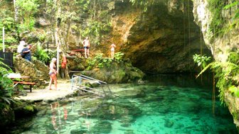 Eine Gruppe von Menschen steht an einem klaren, türkisfarbenen Wasser in einer natürlichen Höhle, umgeben von üppigem Grün.
