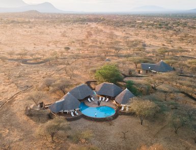 Eine Luftaufnahme eines luxuriösen Safari-Camps mit einem Pool, umgeben von trockener Savanne und sanften Hügeln im Hintergrund.
