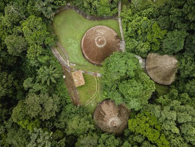 Eine Vogelperspektive zeigt eine üppige Landschaft mit runden Hütten, umgeben von dichtem, grünen Regenwald.