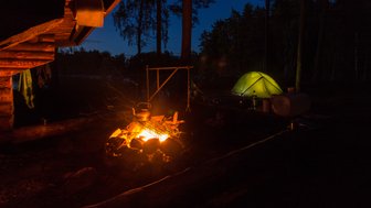 Ein leuchtendes Lagerfeuer erhellt die Dunkelheit, während ein grünes Zelt im Hintergrund steht und Bäume umgeben.