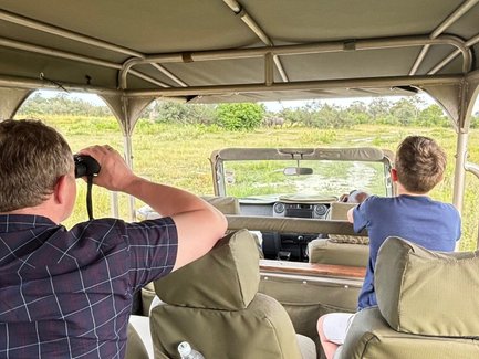 Zwei Personen sitzen in einem Geländewagen und beobachten die Natur, während ein Elefant im Hintergrund sichtbar ist.