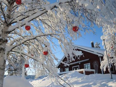 Ein verschneiter Baum mit roten Weihnachtskugeln vor einer rustikalen Hütte in einer winterlichen Landschaft.