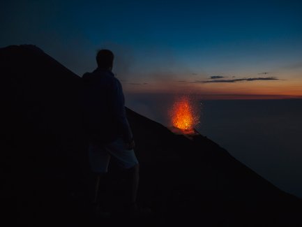 Ein Mensch steht am Rand eines Vulkans und beobachtet die beeindruckende Eruption mit glühenden Lavafontänen im Abendlicht.