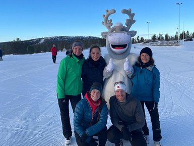 Eine Gruppe von fünf Personen posiert fröhlich im Schnee, umgeben von einer winterlichen Landschaft und einem Rentier-Maskottchen.