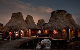 Die strohgedeckten Hütten des Resorts strahlen in der Dämmerung, während ein Boot am Steg festgemacht ist.