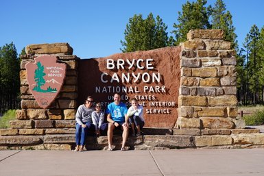 Eine Familie sitzt fröhlich vor dem Eingangsschild des Bryce Canyon Nationalparks, umgeben von Bäumen und klarem Himmel.