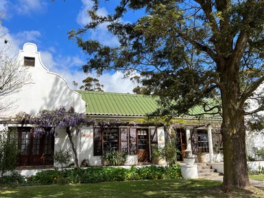 Ein charmantes Gästehaus mit einem grünen Dach, umgeben von blühenden Pflanzen und einem großen Baum im Vordergrund.