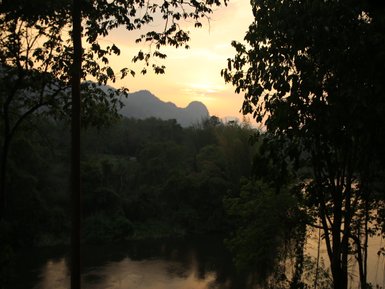 Ein ruhiger Fluss fließt sanft durch eine grüne Landschaft, umgeben von Bergen und Bäumen im warmen Licht der Dämmerung.